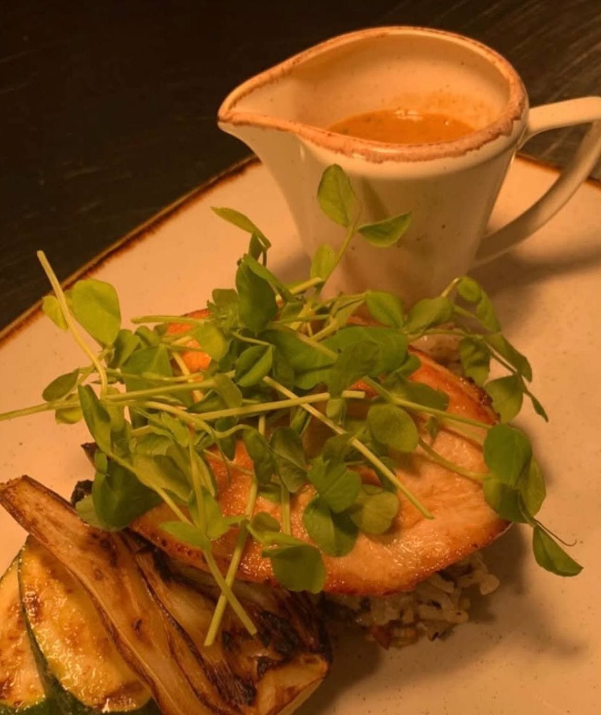 Salmon on rice with charred vegetables, topped with fresh pea shoots, served with a jug of sauce on the side.