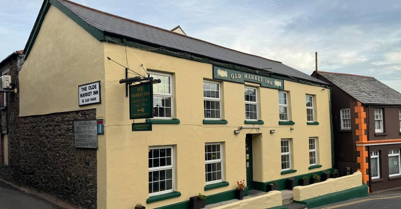 The Olde Market Inn on Chapel Street, Holsworthy -- cream-rendered frontage with green trim, 'OLD MARKET INN' signboards and 'Good Food · Fine Ales' hanging sign.