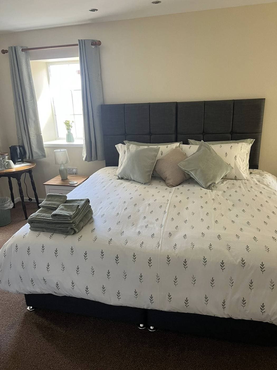 An en-suite letting room above the pub -- king-sized bed with a grey headboard, leaf-pattern bedding, natural light from a sash window and fresh towels folded on the bed.